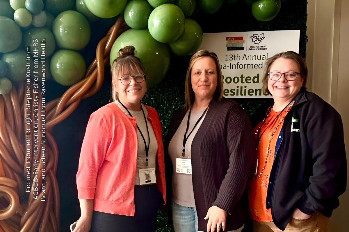 Three women smiling together in front of a green balloon tree backdrop at the Ohio Department of Behavioral Health's 13th Annual Trauma-Informed Conference: Rooted in Resilience.